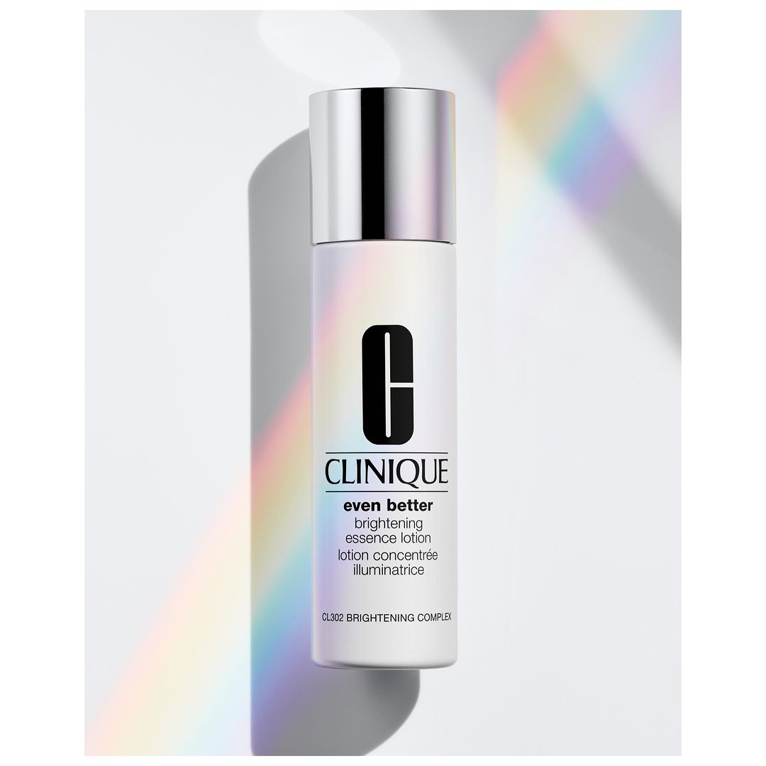 Close-up of a Clinique Even Better brightening essence lotion bottle with a silver cap on a white surface with pastel light.