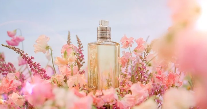 Close-up of a perfume bottle surrounded by pink and white flowers, set against a pale sky, creating a soft, floral fragrance visual.