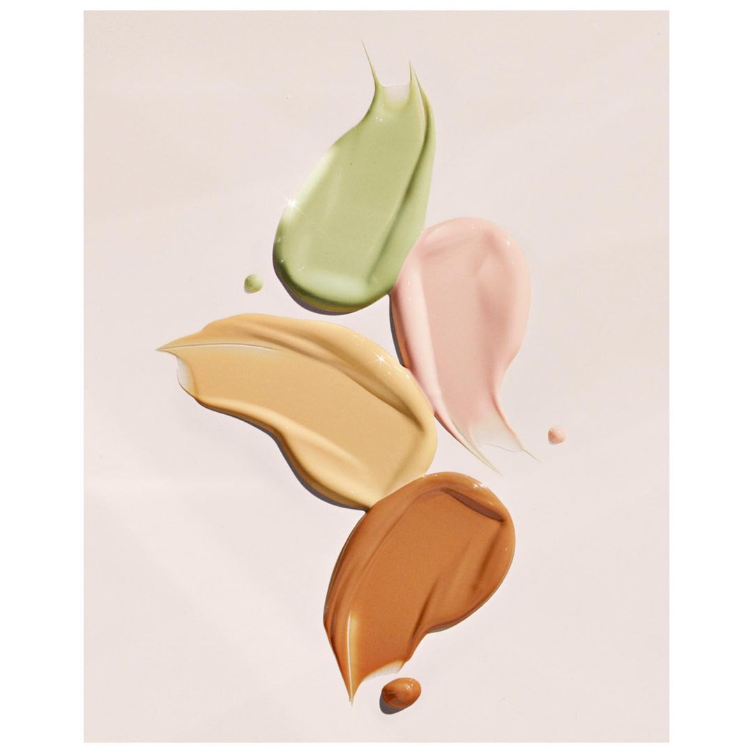 Swatches of various shades of liquid makeup, including green, tan, peach, and brown, are displayed on a white background.