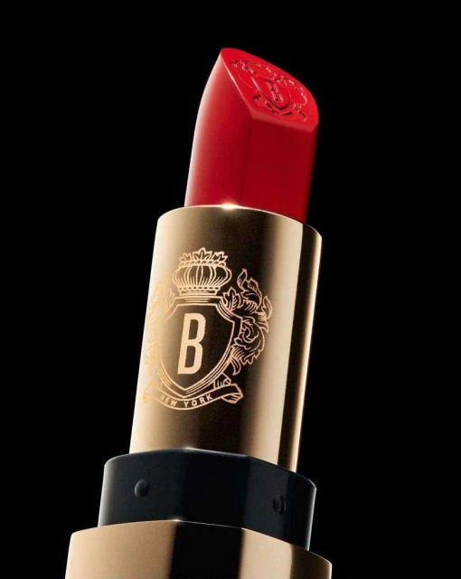 A close-up studio shot of a vibrant red lipstick with gold casing and an ornate logo against a stark black background, beauty product.