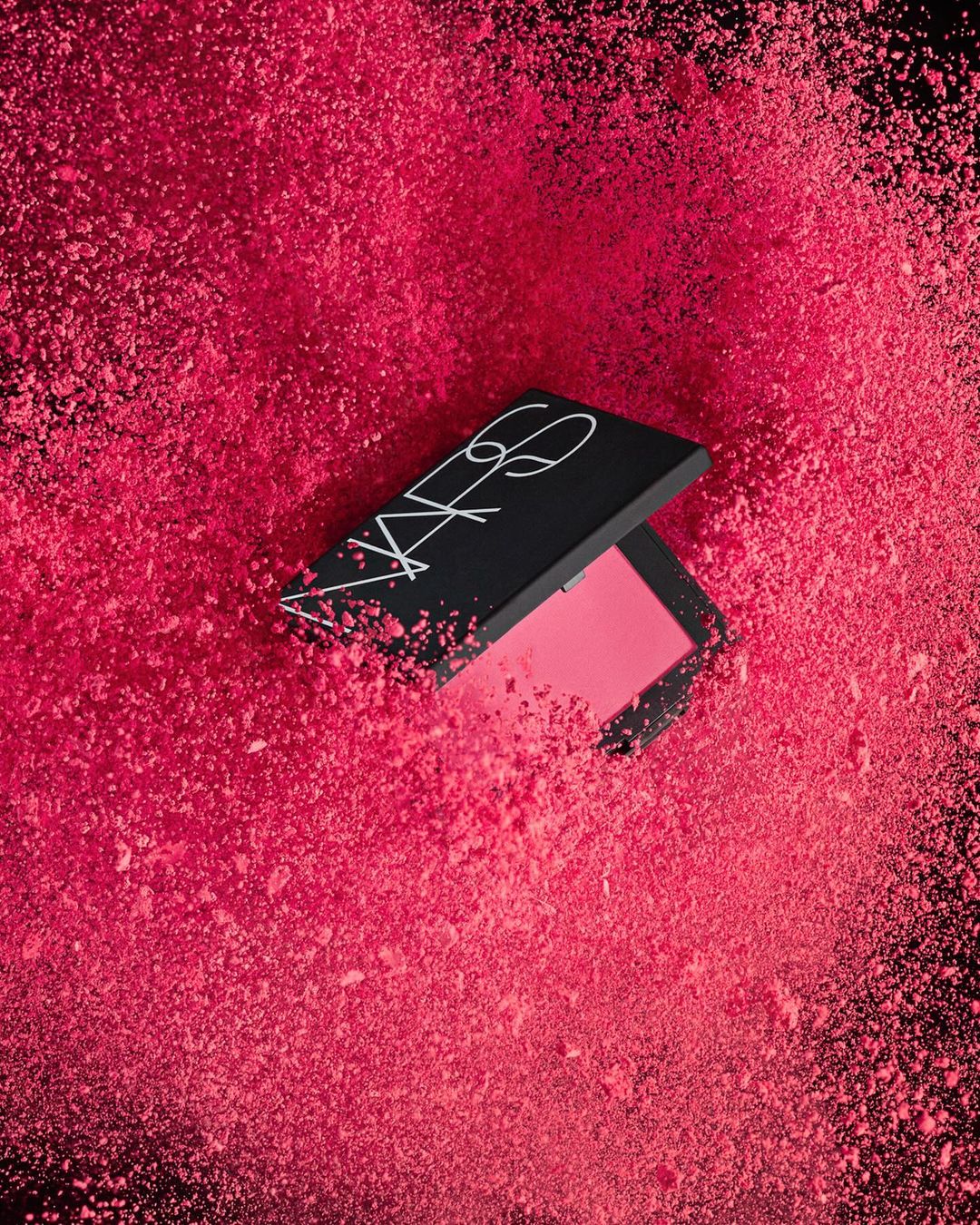 NARS blush product surrounded by a cloud of pink powder against a black background in a beauty or fashion-related image.