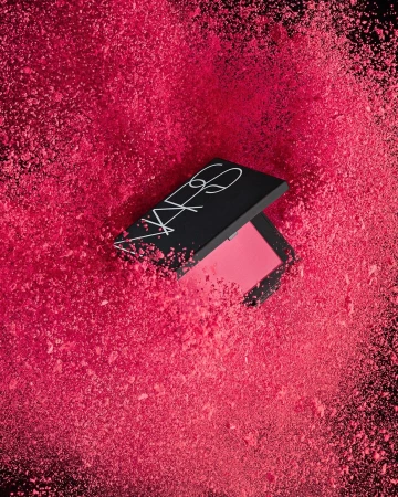 NARS blush product surrounded by a cloud of pink powder against a black background in a beauty or fashion-related image.