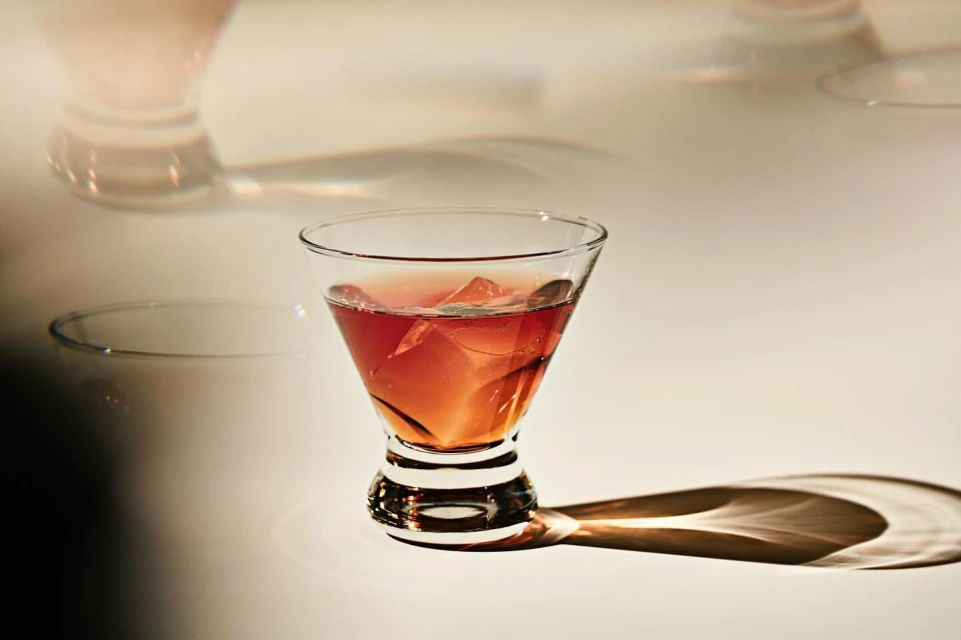 Crystal glass containing a red cocktail with ice cubes on a reflective surface, casting shadows in a warm-toned ambiance.