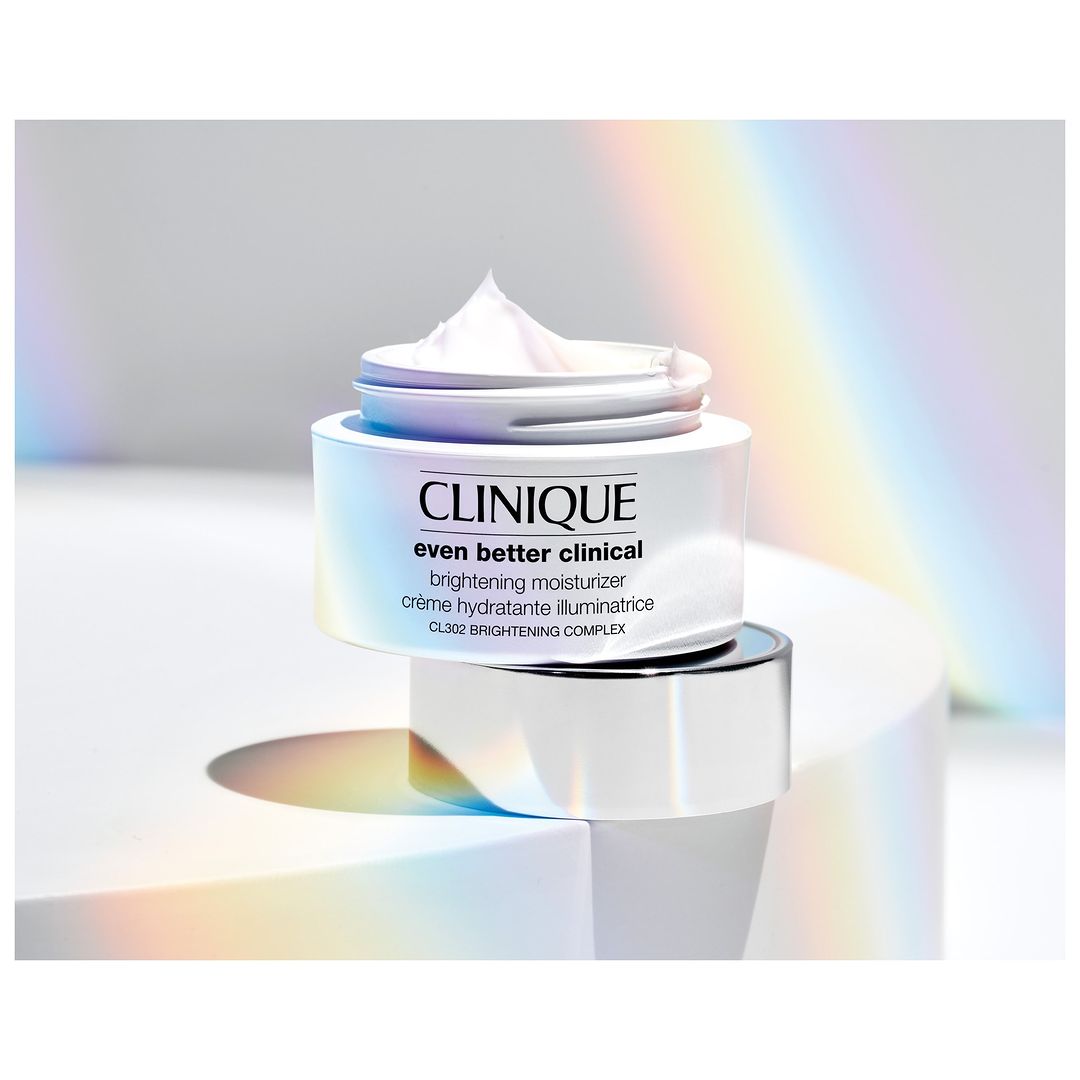 Clinique 'Even Better Clinical' brightening moisturizer on a white podium against a soft, rainbow-lit background in a beauty still life.