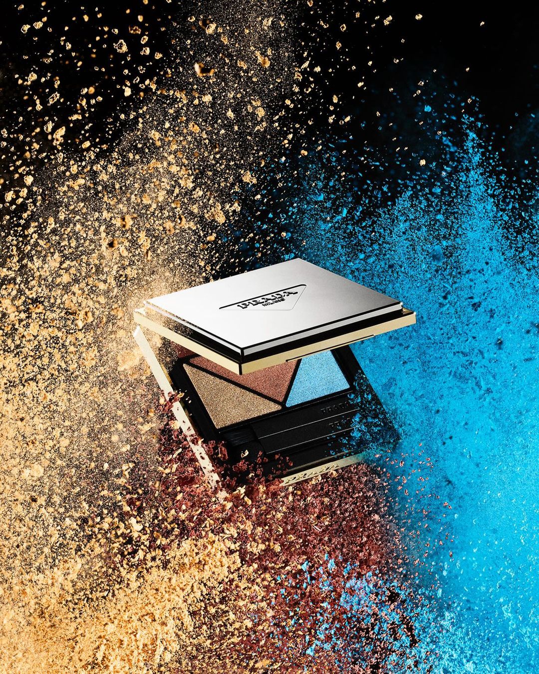 Eye shadow palette surrounded by blue and bronze powder explosion against a black backdrop in a beauty or cosmetics shoot.
