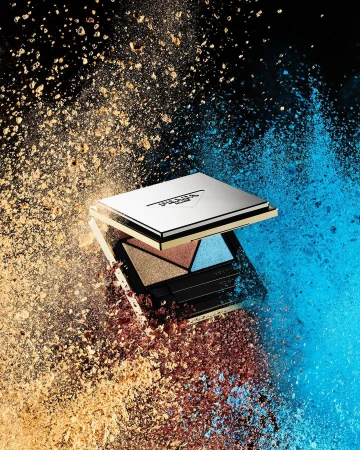Eye shadow palette surrounded by blue and bronze powder explosion against a black backdrop in a beauty or cosmetics shoot.