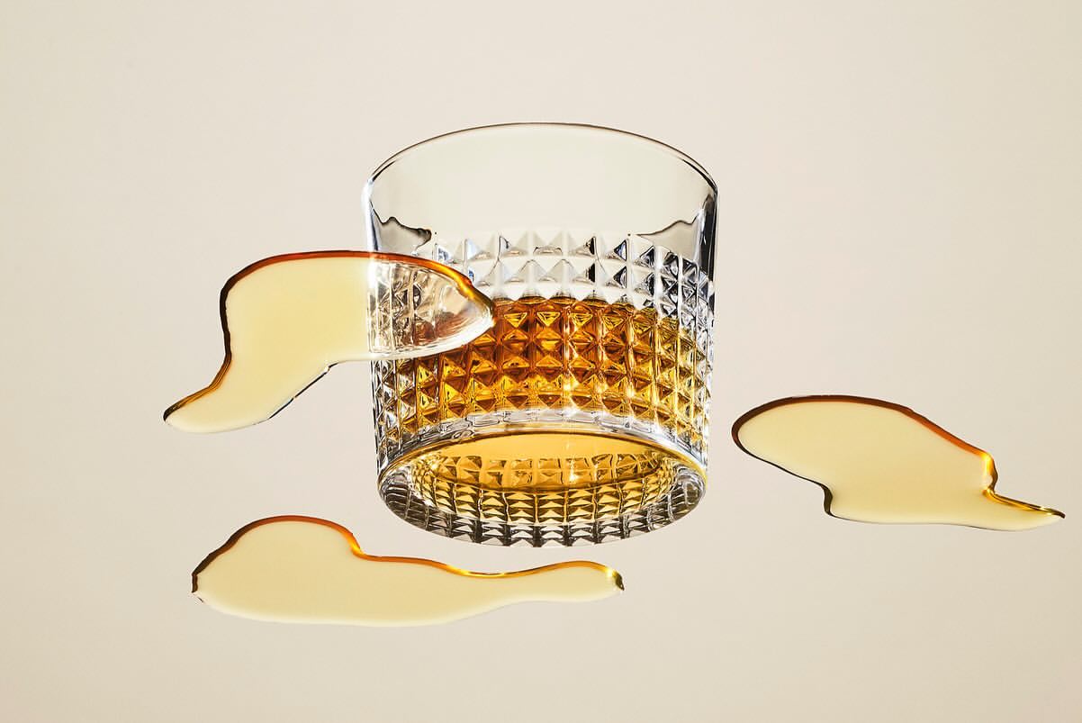 Crystal whiskey glass filled with amber liquid, accompanied by spills on a beige surface. Drink photography for the food and beverage industry.
