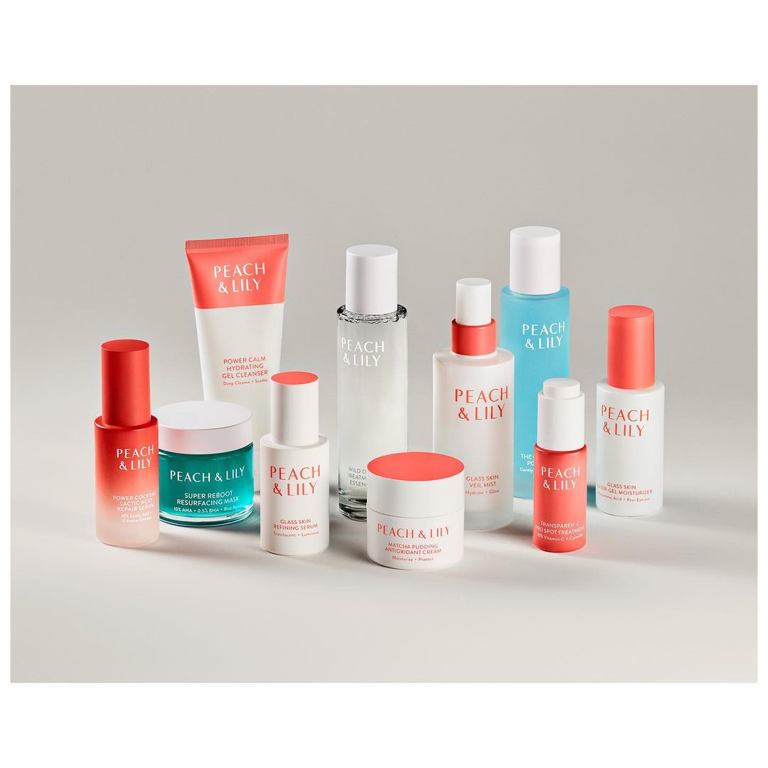 Display of Peach & Lily skincare products featuring bottles, tubes, and jars against a soft gray background in a studio setting.
