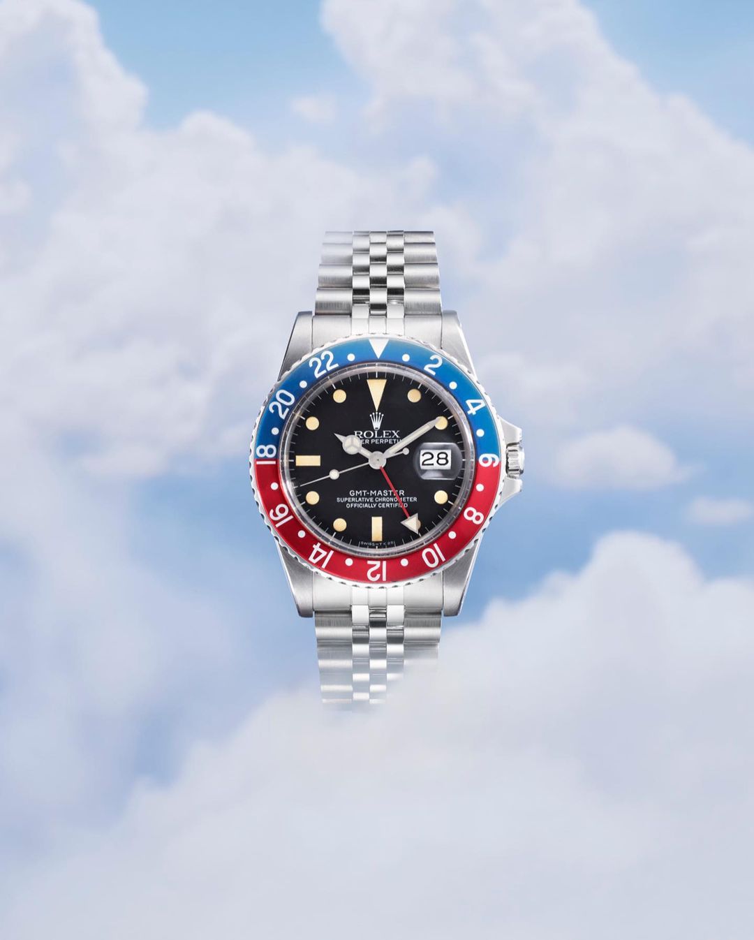 Wristwatch with a jubilee bracelet, black dial, and Pepsi bezel against a blurred sky background with clouds, captured in a studio shot.