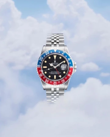 Wristwatch with a jubilee bracelet, black dial, and Pepsi bezel against a blurred sky background with clouds, captured in a studio shot.