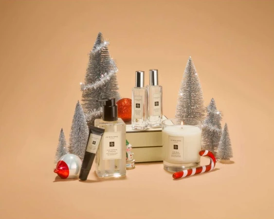 Jo Malone London products displayed with miniature Christmas trees, ornaments, a candle, and a candy cane on a light brown backdrop.