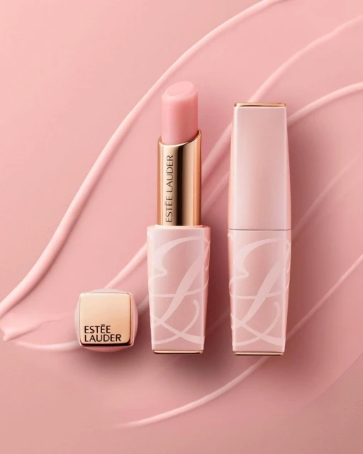 Three tubes of Estee Lauder lipstick are displayed on a pink surface with abstract pink smears. One tube is open, showing the lipstick.