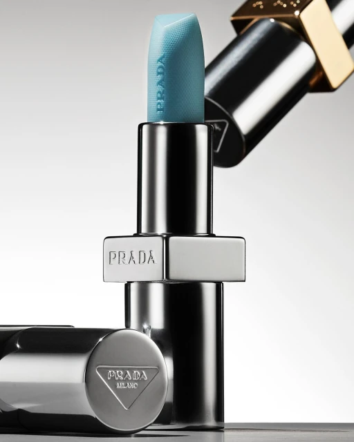 Close-up studio shot of teal Prada lipstick with chrome and gold hardware against a gradient gray background. Beauty and fashion.