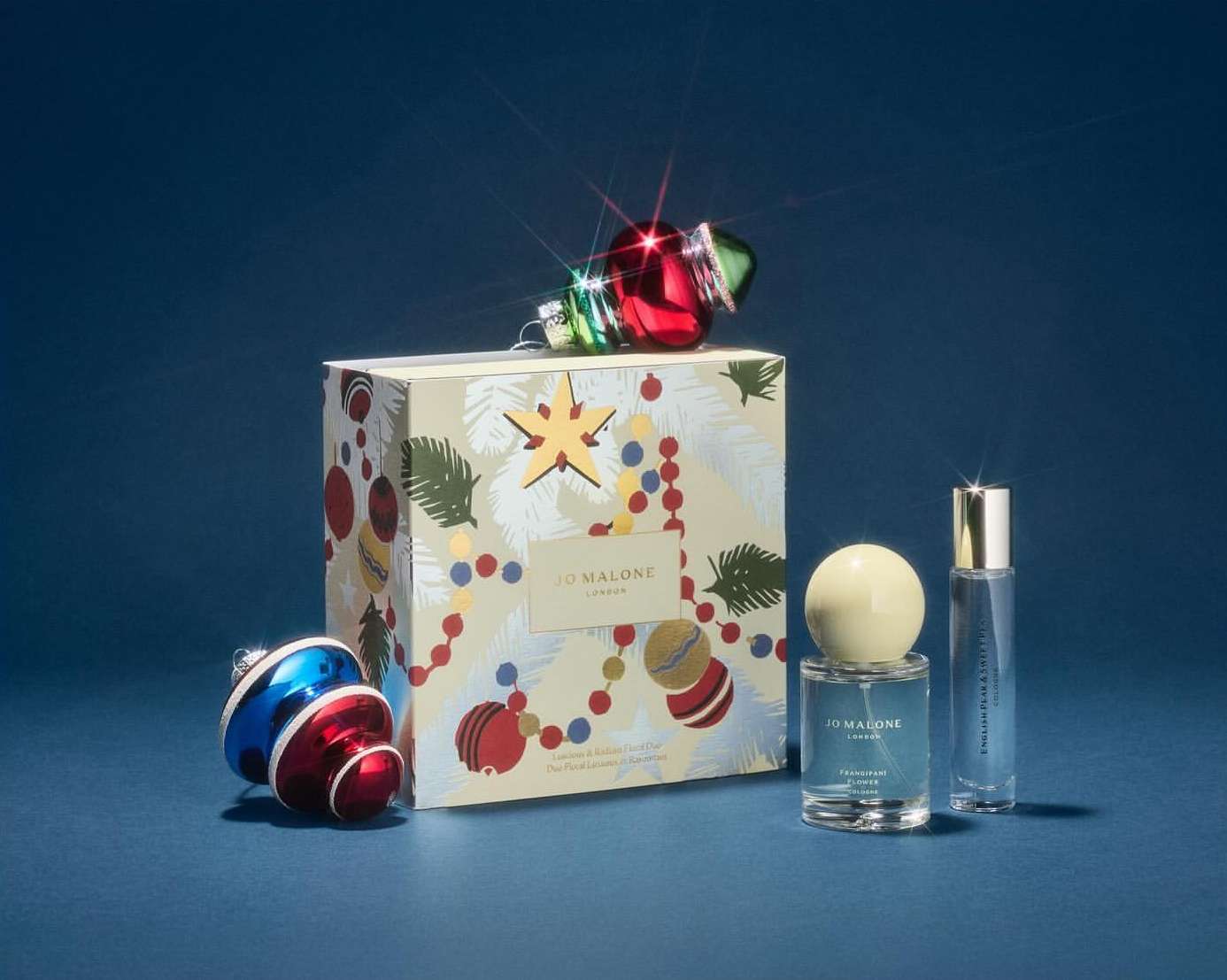 Jo Malone holiday gift set featuring perfumes, seasonal packaging with Christmas ornaments, on a blue backdrop with shiny reflections.