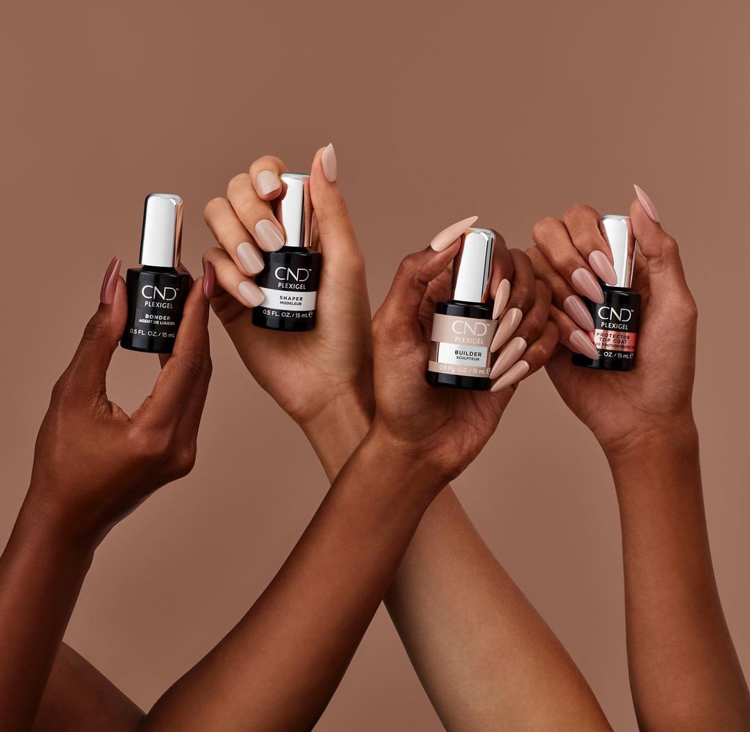 Four diverse hands holding CND Plexigel Builder nail polish bottles in neutral shades against a warm, tan backdrop in a beauty studio setting.