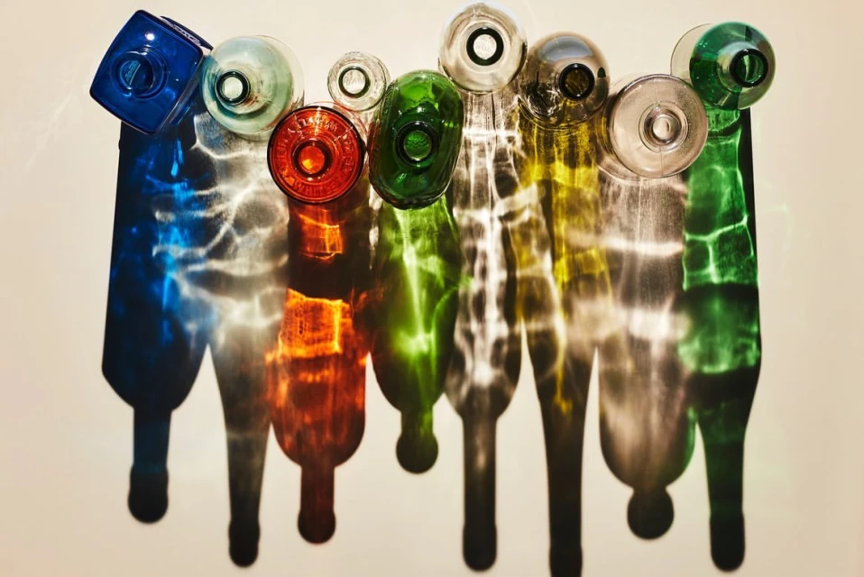 Colorful glass bottles in a row cast long, vibrant shadows on a cream surface, highlighting their forms and hues in a minimalist scene.