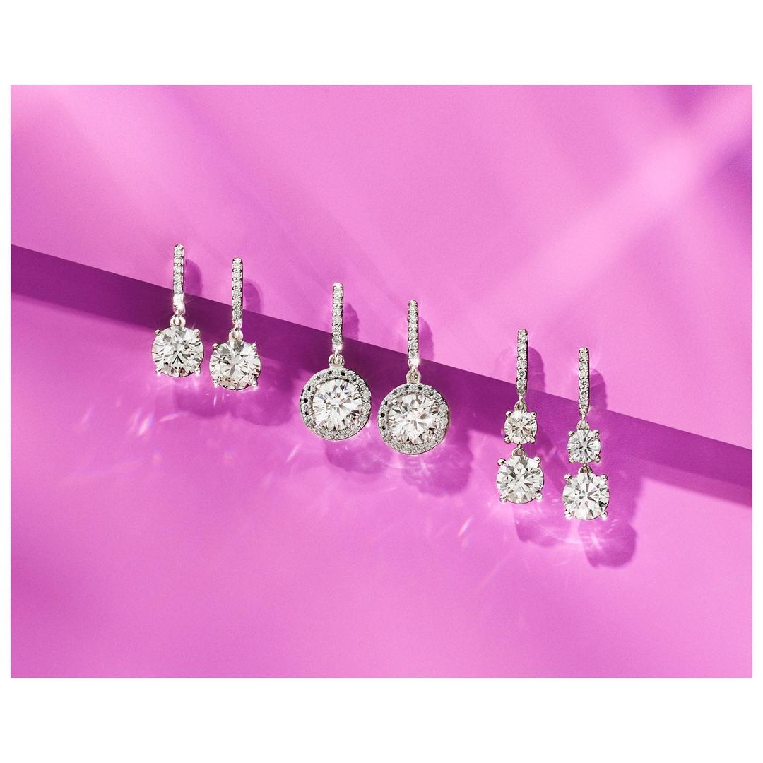 Three pairs of diamond drop earrings with varying stone shapes and halo settings on a bright purple background, jewellery photography.