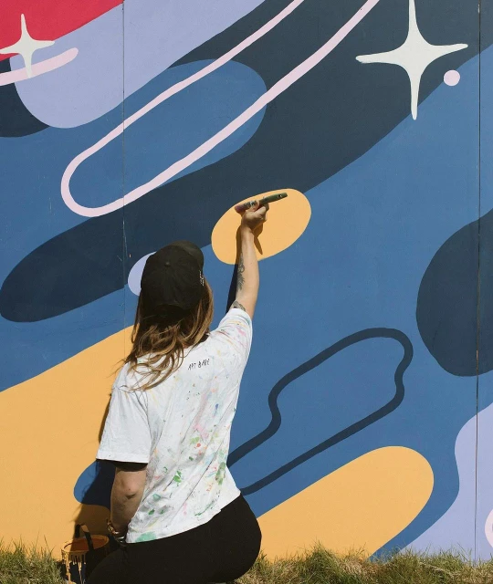 Artist paints colorful mural on wall outdoors wearing a cap and paint-splattered white t-shirt, with abstract design and stars.