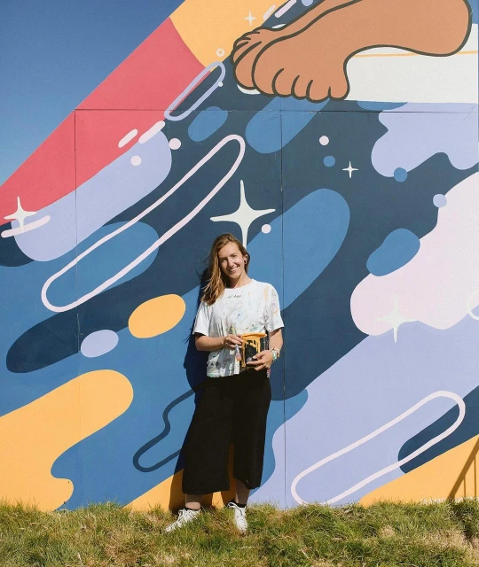 Smiling woman artist stands by colorful mural on bright day, holding a box, wearing a white t-shirt and black culottes.