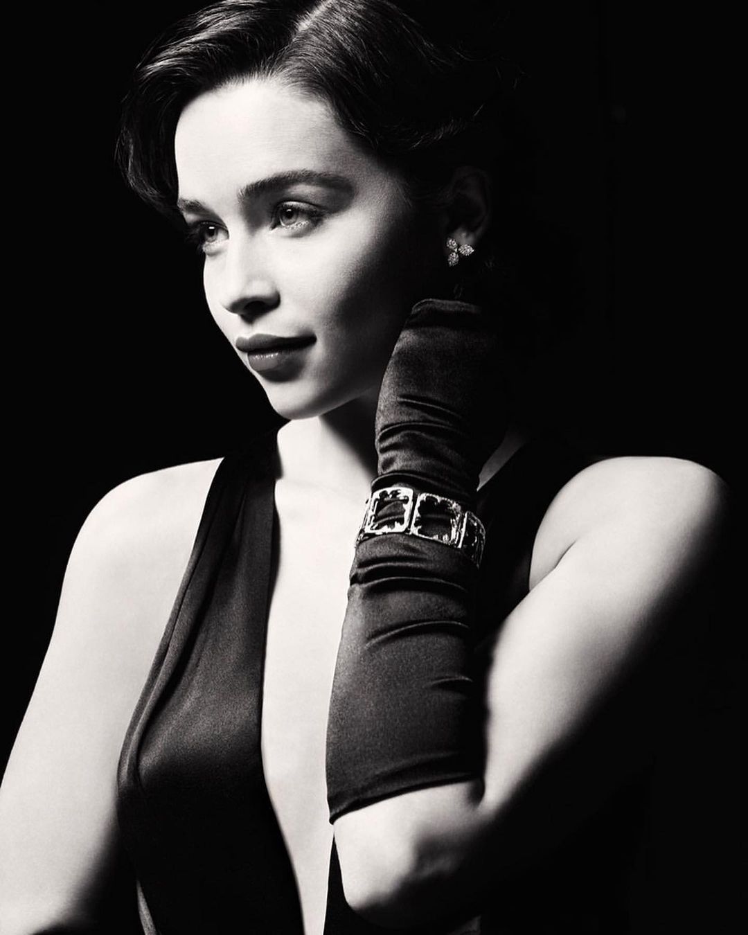 Portrait of a woman with dark hair wearing an elegant sleeveless dress, long gloves, and a bracelet, posing against a black backdrop.