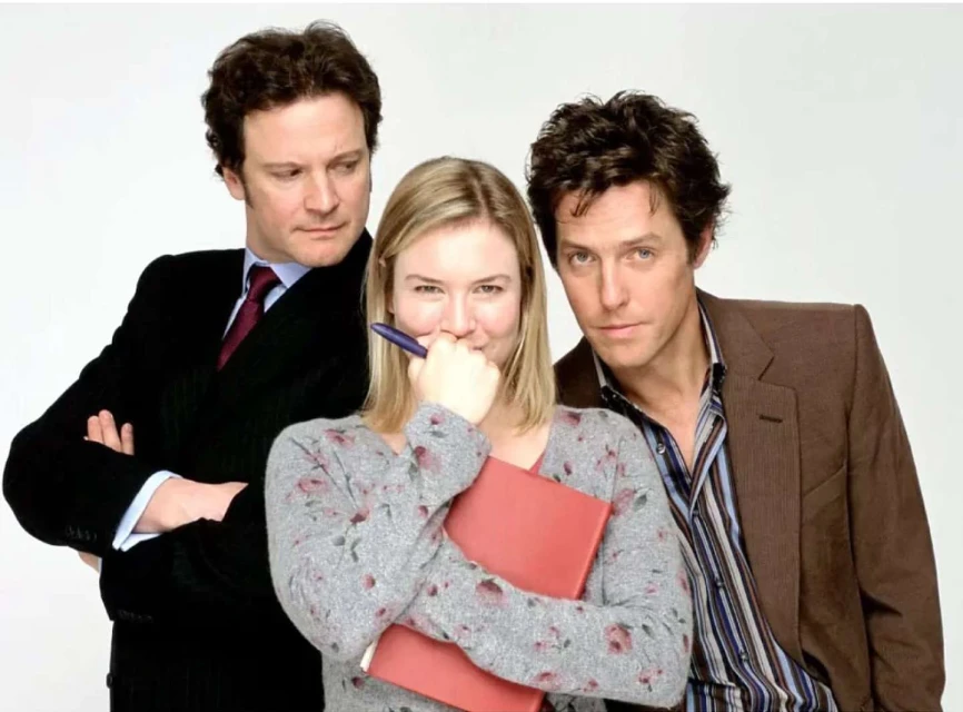 Three actors pose together in a promotional shot. A woman in the center holds a pen and folder, flanked by two men in suits.