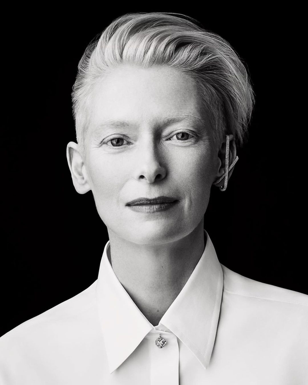 Black and white portrait of Tilda Swinton in white collared shirt with short platinum hair, earring, against a black backdrop.