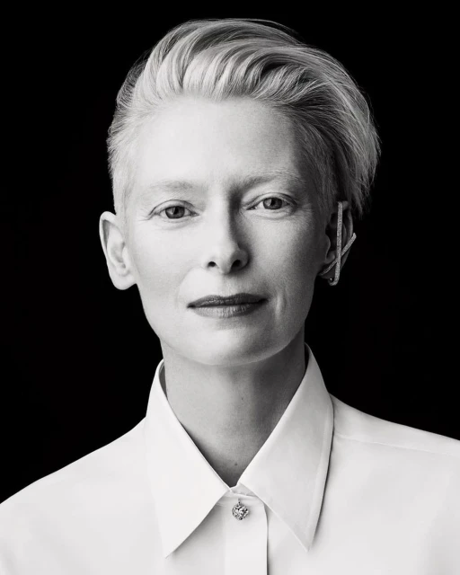 Black and white portrait of Tilda Swinton in white collared shirt with short platinum hair, earring, against a black backdrop.