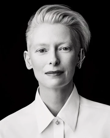Black and white portrait of Tilda Swinton in white collared shirt with short platinum hair, earring, against a black backdrop.