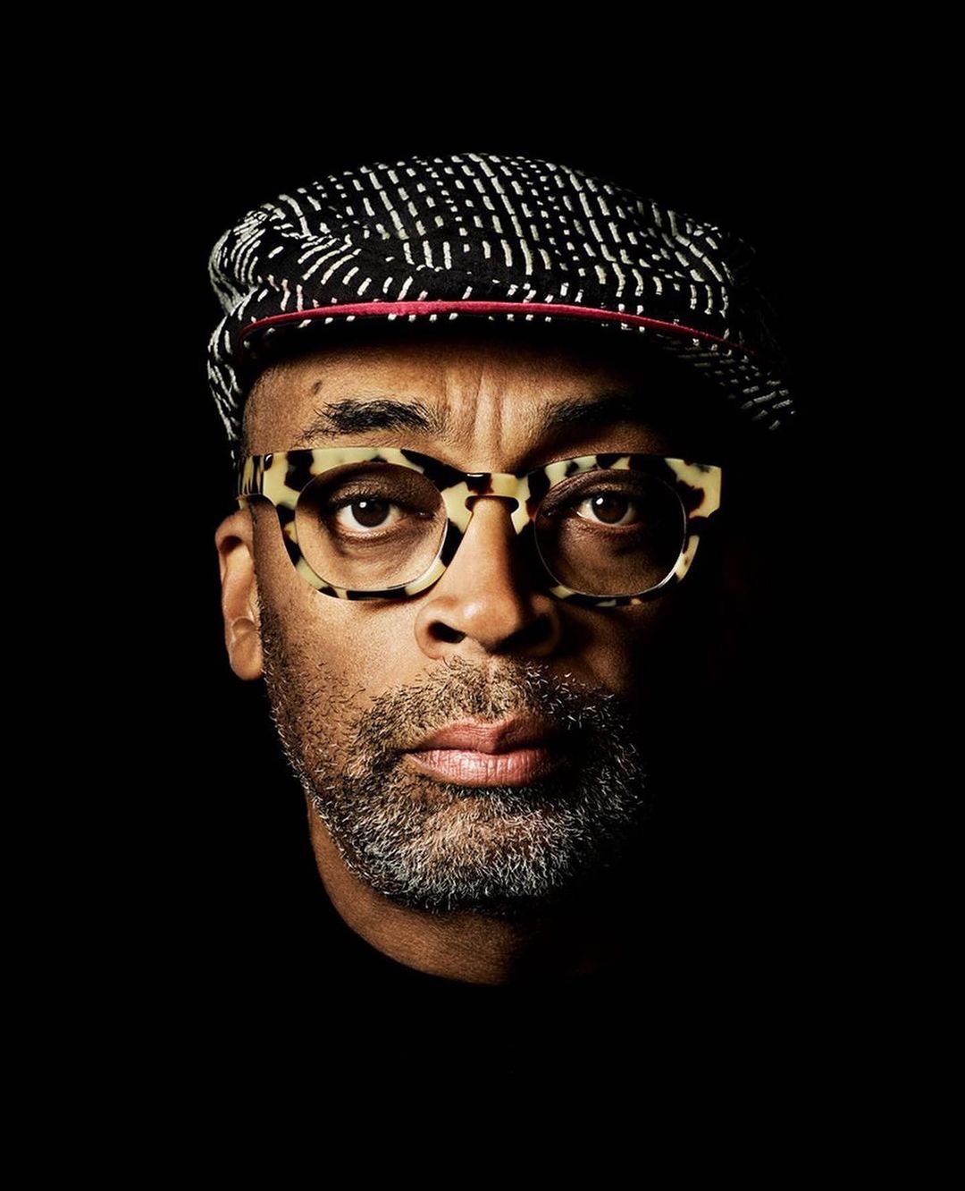 Close-up portrait of a stylish adult male with patterned cap and tortoiseshell glasses against a dark background, showcasing fashion and personality.