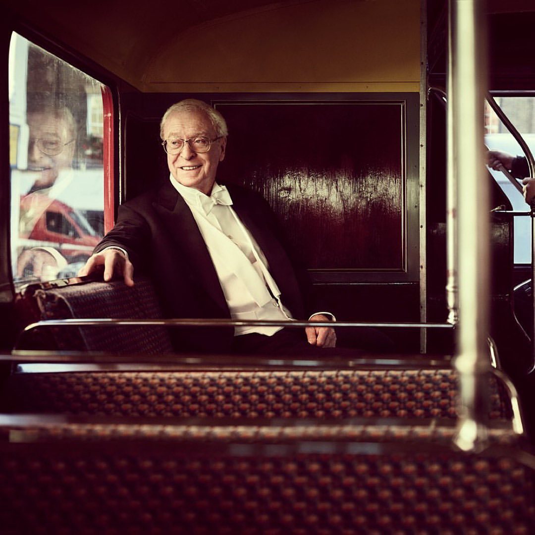 Elderly man in a black tuxedo and white tie smiles inside a vintage bus with patterned seating and warm interior lighting.