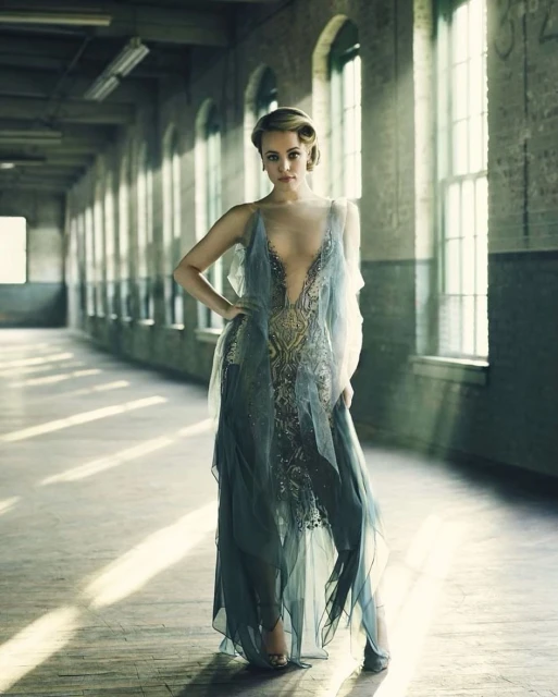 Elegant woman in sheer blue gown strikes pose in an industrial-style corridor with large windows, bathed in soft, directional light.