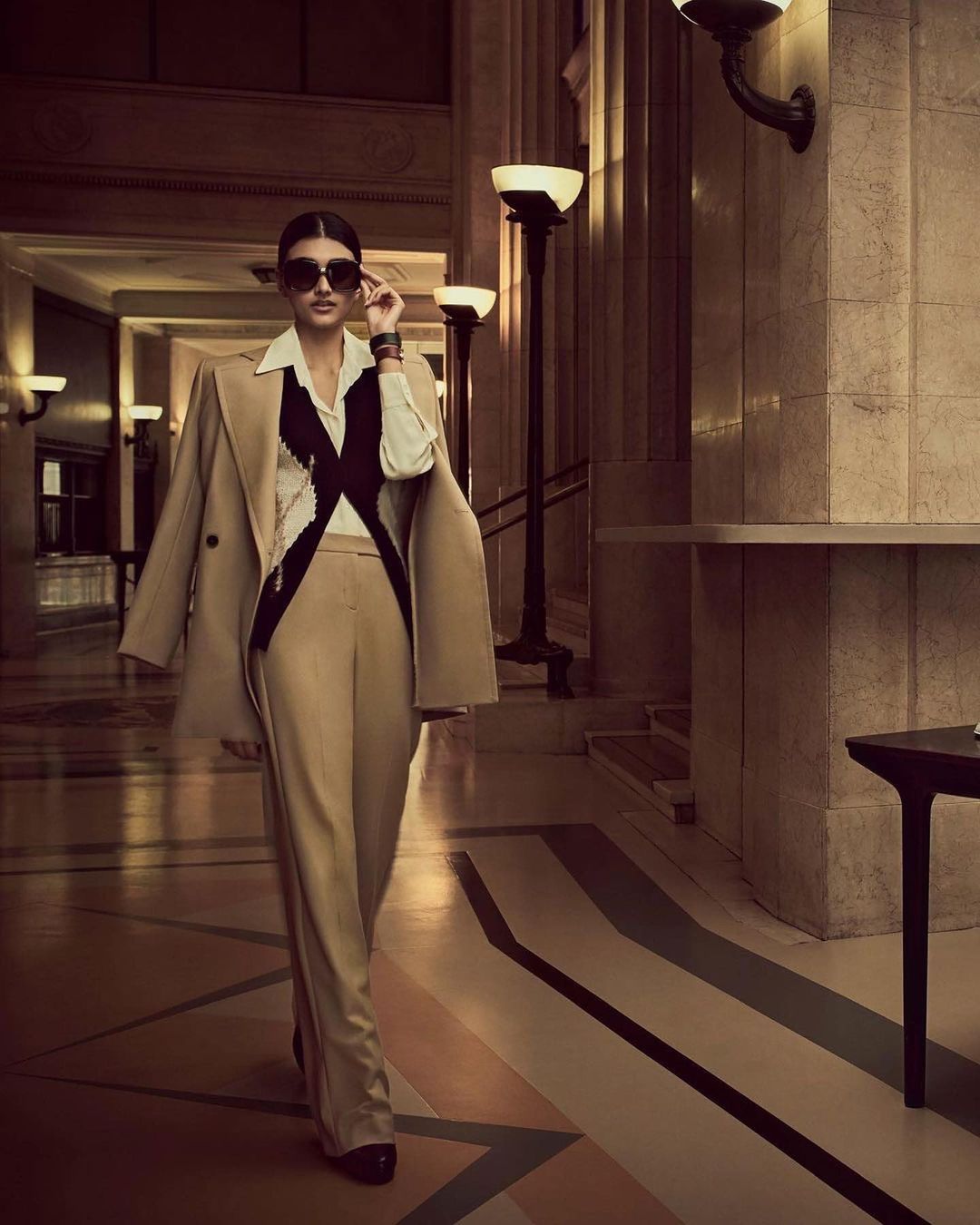 Woman in a tan suit and sunglasses walking in a building with marble columns and vintage lighting fixtures, fashion editorial style.