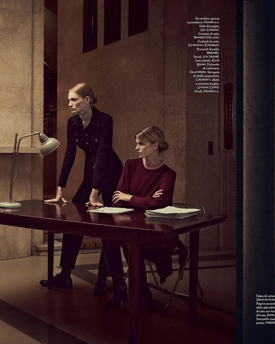 Two women in formal wear pose at a desk indoors with a green lamp, papers, and dark wood furniture in a dimly lit setting.