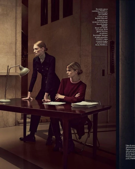 Two women in formal wear pose at a desk indoors with a green lamp, papers, and dark wood furniture in a dimly lit setting.