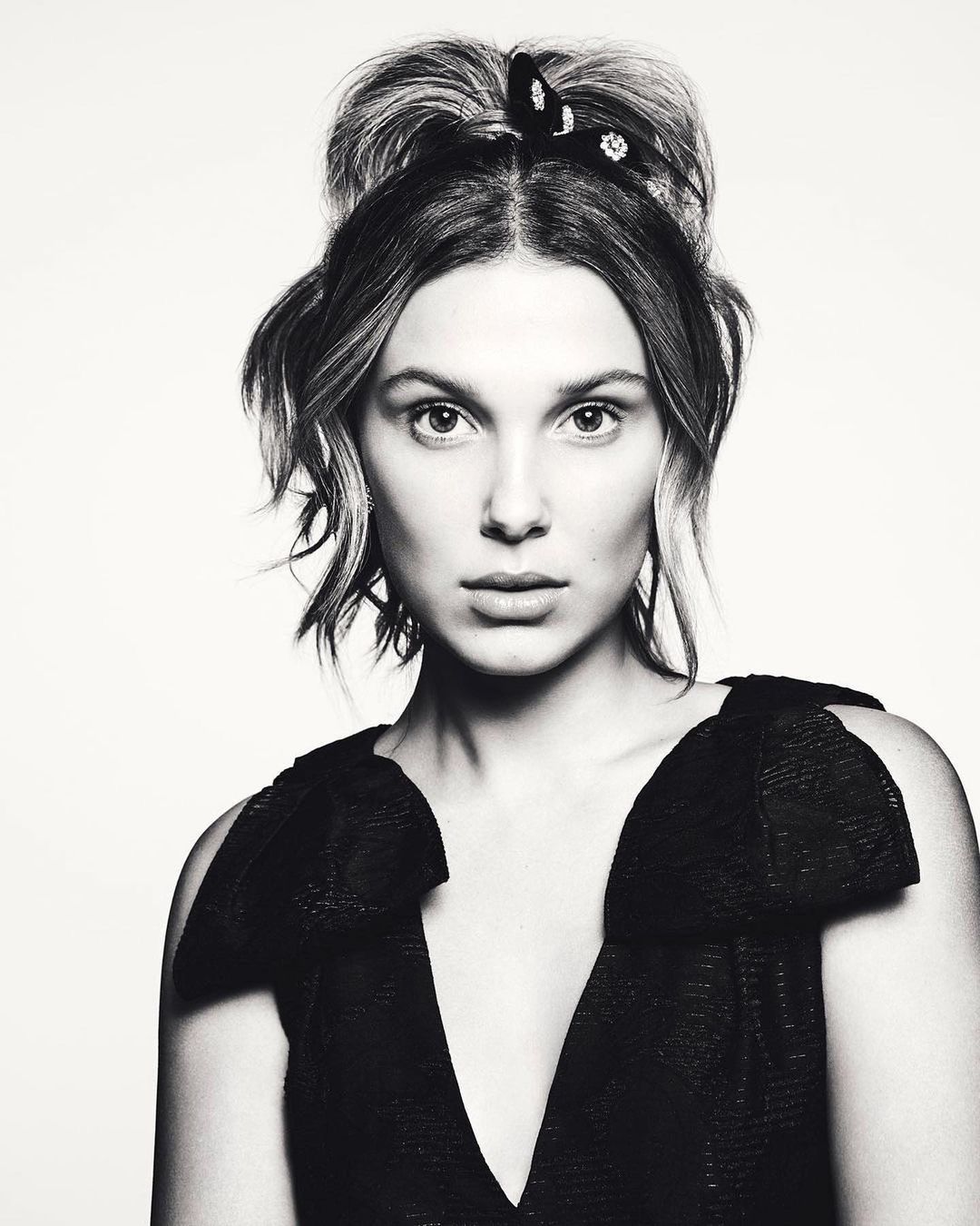 Black and white portrait of a young woman with her hair up, wearing a black dress with a V-neck, against a white background.