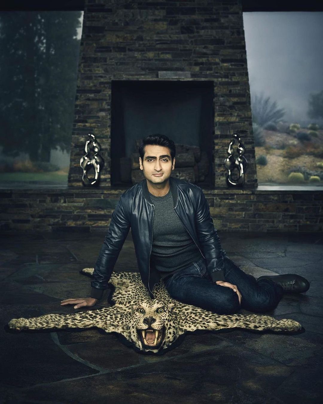 Portrait of Kumail Nanjiani in a leather jacket sitting on a jaguar-print rug, with a modern fireplace and nature visible in the background.