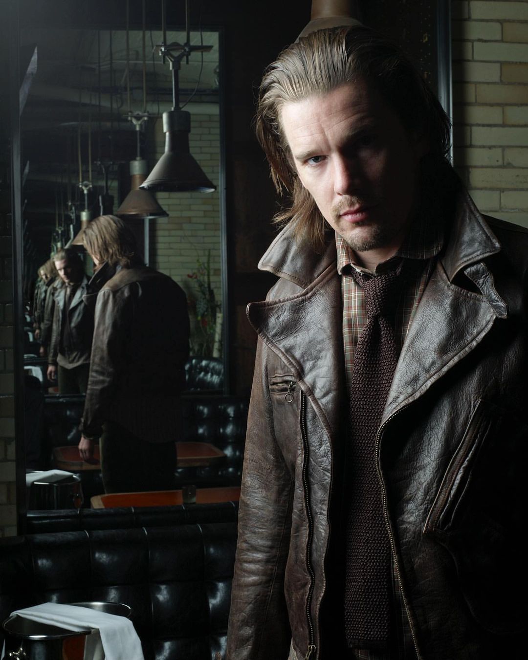 Portrait of an adult man with long hair wearing a brown leather coat in a dimly lit interior with mirrors and vintage decor.