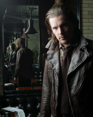 Portrait of an adult man with long hair wearing a brown leather coat in a dimly lit interior with mirrors and vintage decor.
