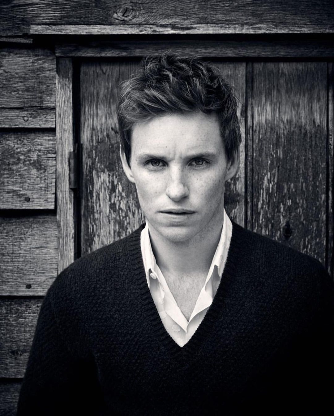 Black and white portrait of a young adult male with fair skin and short brown hair, wearing a v-neck sweater and white shirt, against a wooden wall.