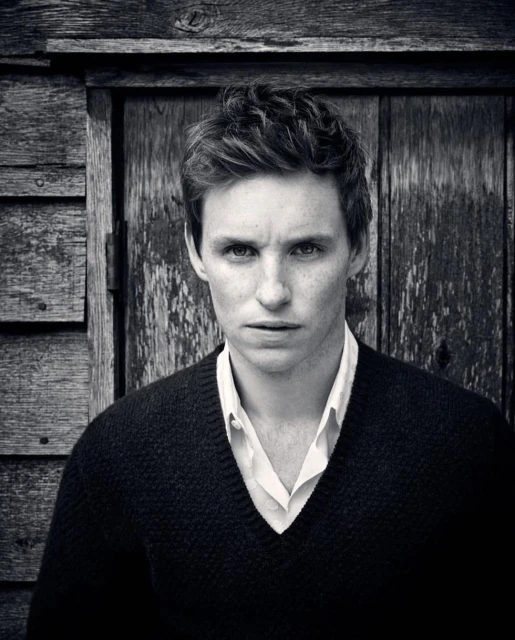 Black and white portrait of a young adult male with fair skin and short brown hair, wearing a v-neck sweater and white shirt, against a wooden wall.