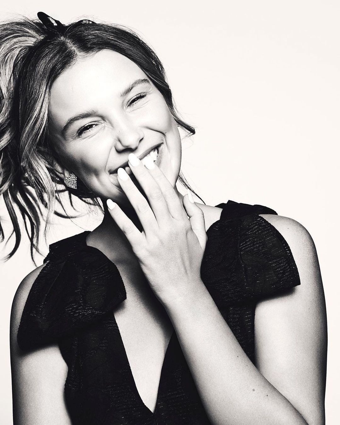Monochrome portrait of a smiling woman with her hand near her face, wearing a V-neck top with shoulder bows, captured in an indoor setting.