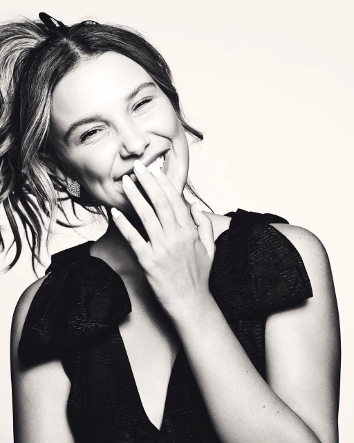 Monochrome portrait of a smiling woman with her hand near her face, wearing a V-neck top with shoulder bows, captured in an indoor setting.