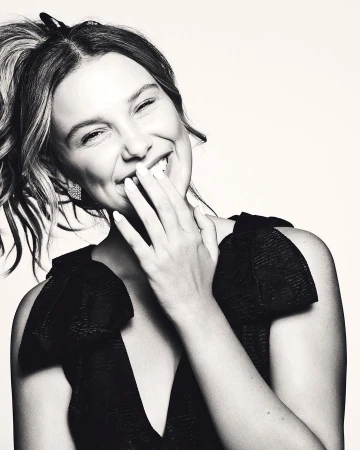 Monochrome portrait of a smiling woman with her hand near her face, wearing a V-neck top with shoulder bows, captured in an indoor setting.