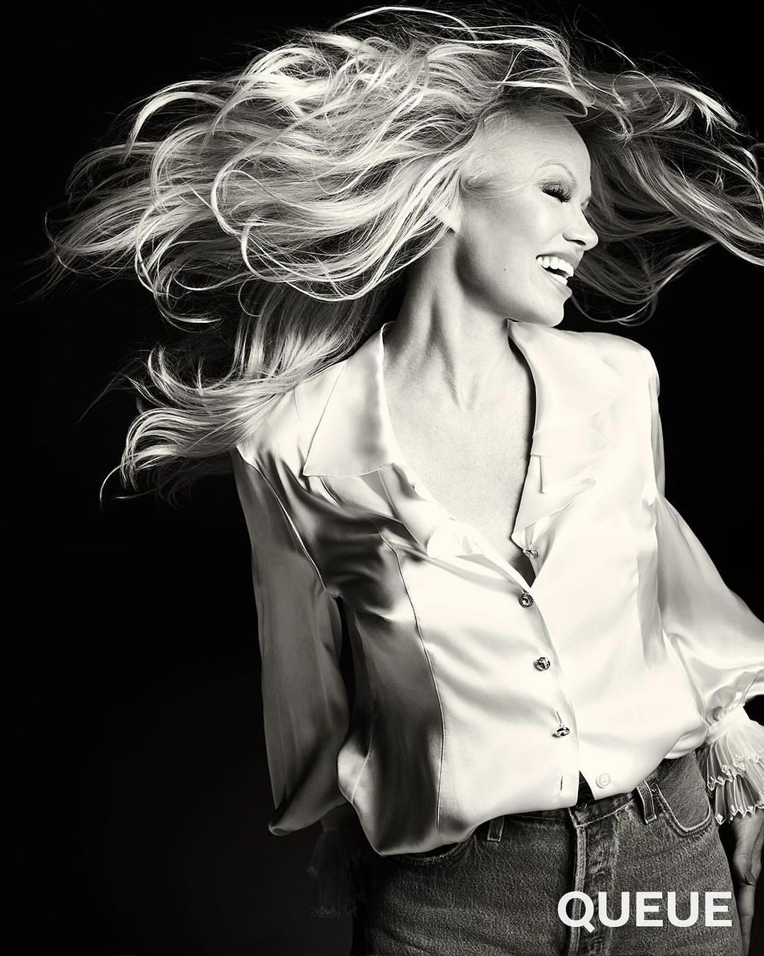 Black-and-white portrait of a smiling woman with long, flowing blonde hair, wearing an unbuttoned satin blouse and jeans.