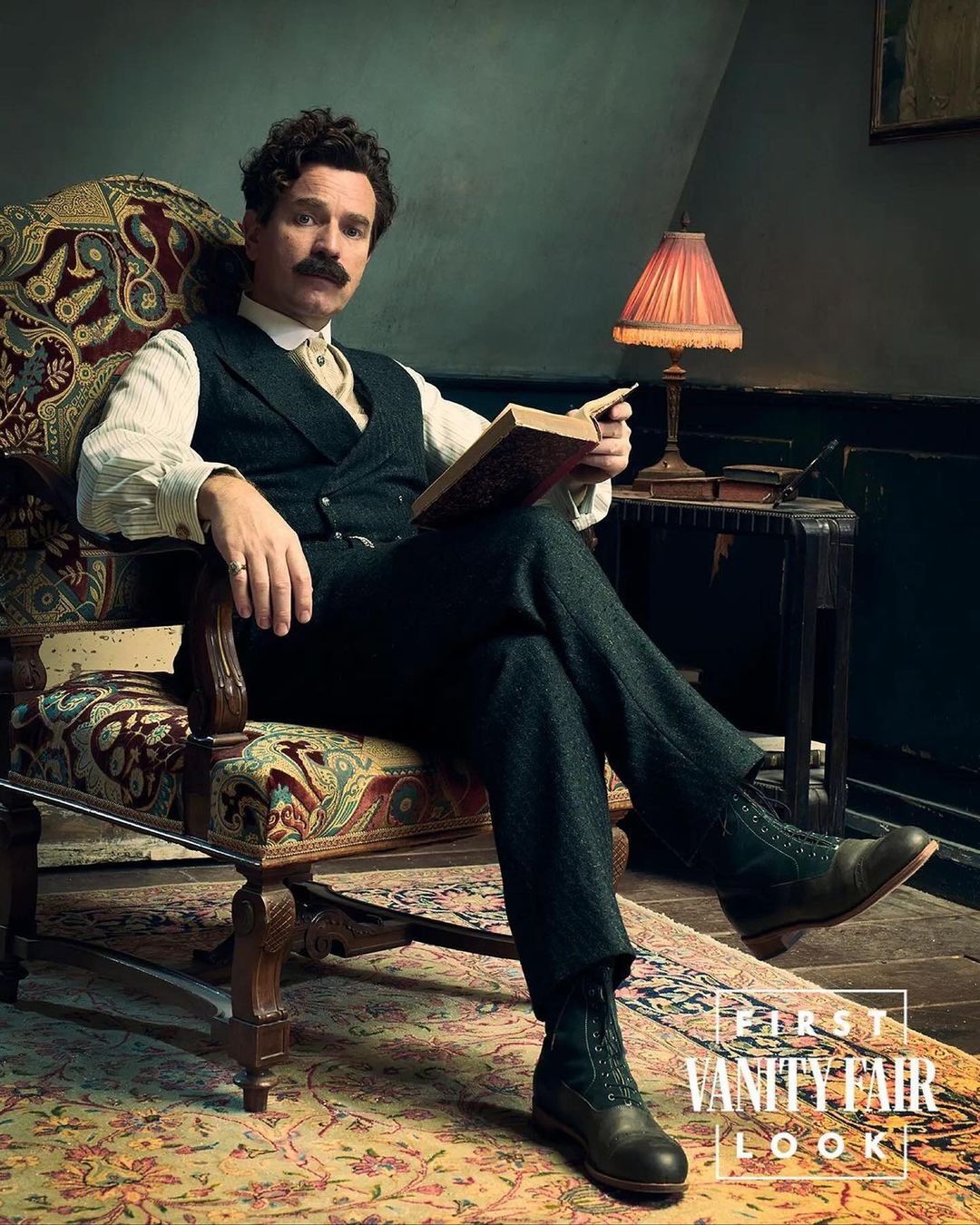 Man with mustache in Victorian-era attire sitting in armchair, reading book indoors with warm lamp light, antique rug on the floor.