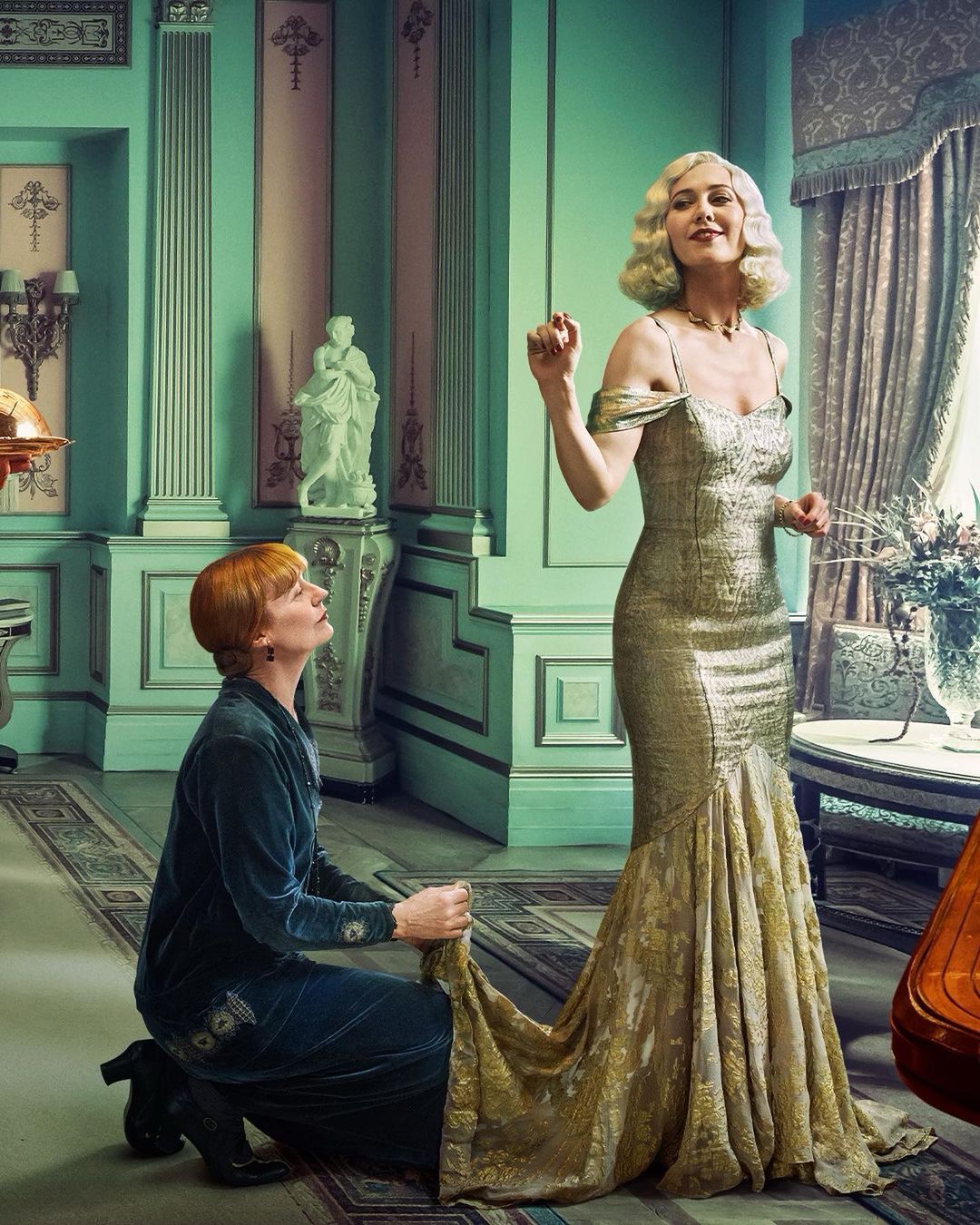 Two women in an ornate room; one in gold gown standing, the other kneeling adjusting the dress's train.