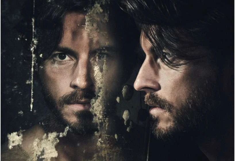 Portrait of an adult man with dark hair and a beard, gazing intently through a distressed mirror, creating a dual perspective.