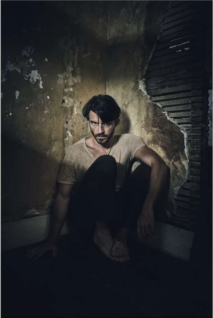 Man sitting on the floor against damaged wall, wearing a t-shirt and dark trousers, looking toward the camera with serious expression.