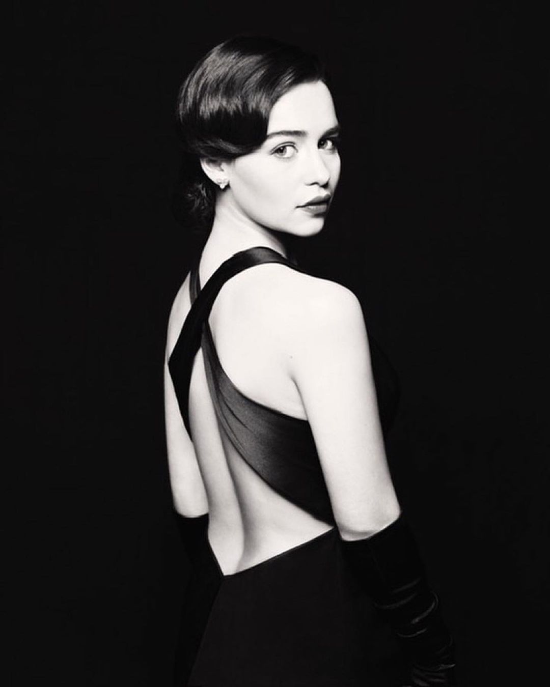 Monochrome portrait of woman in black dress and gloves looking over her shoulder with dark background, short wavy dark hair and red lipstick.