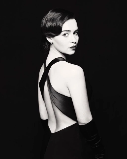 Monochrome portrait of woman in black dress and gloves looking over her shoulder with dark background, short wavy dark hair and red lipstick.